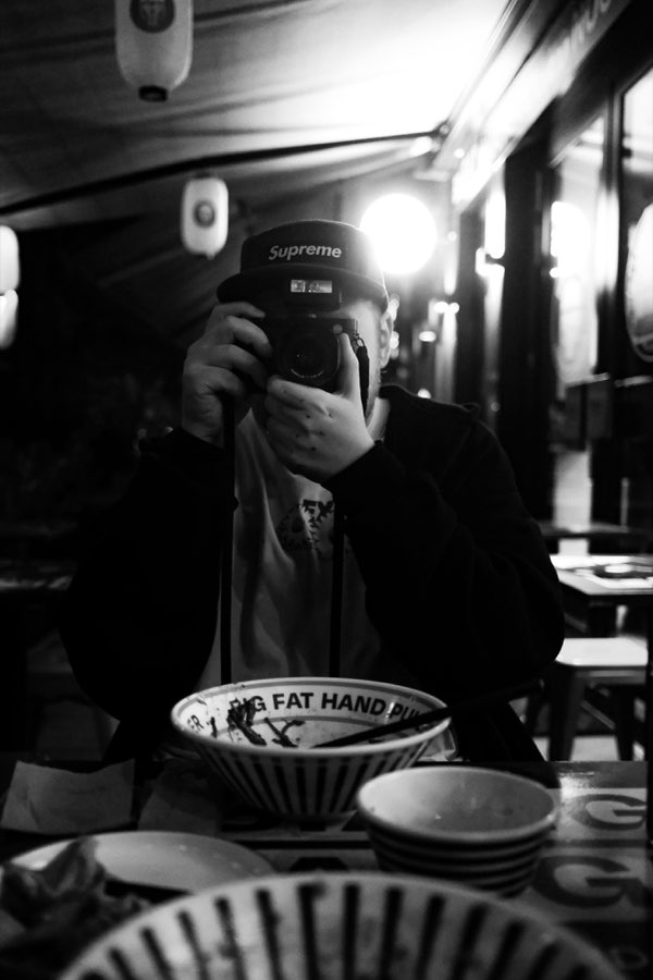 Black and white photo of Julian Aul in a cap, taking a picture with his camera at a restaurant table