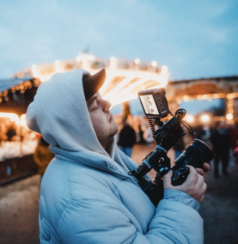 Julian Aul in a hoodie filming with a cinema camera rig at a Christmas market at dusk