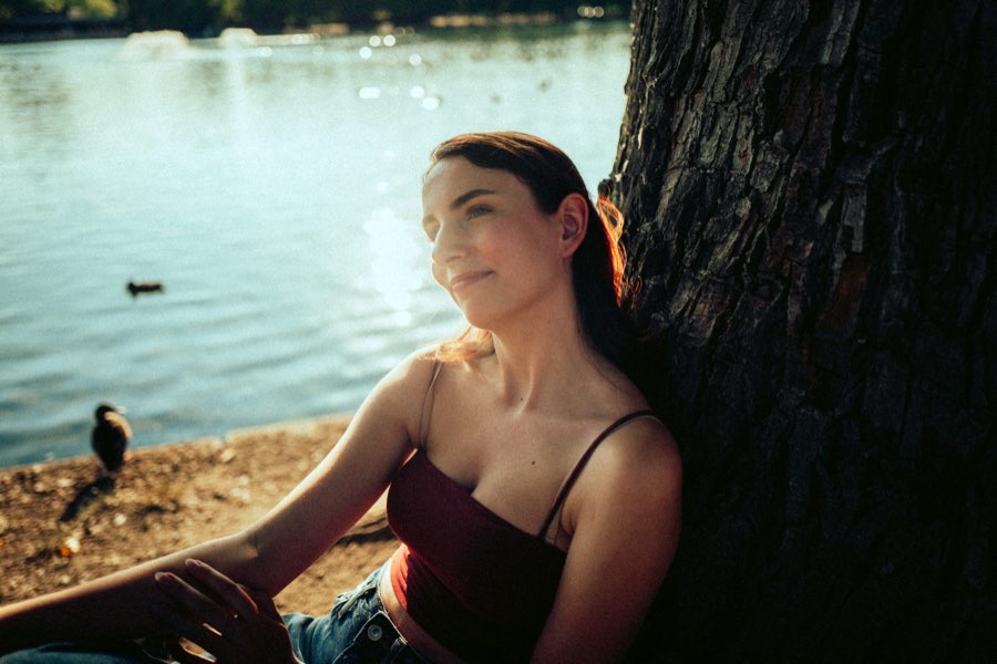 Woman leaning against a tree trunk by a lake in soft sunlight