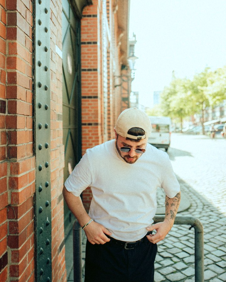 Man in a white T-shirt and cap leaning against a brick wall