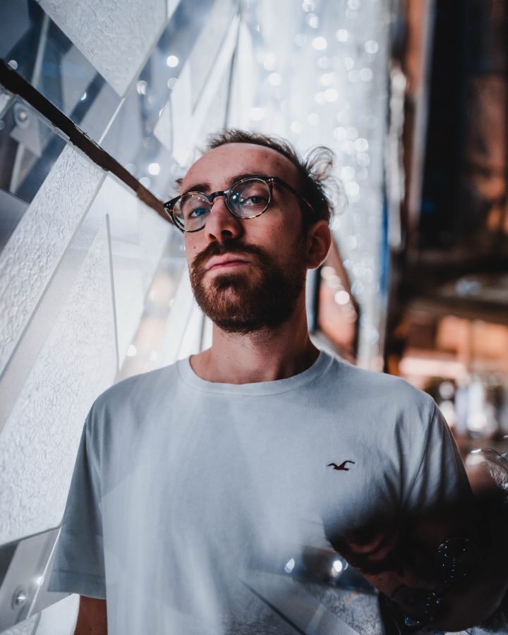 Man with glasses in a grey T-shirt in a dimly lit glass corridor