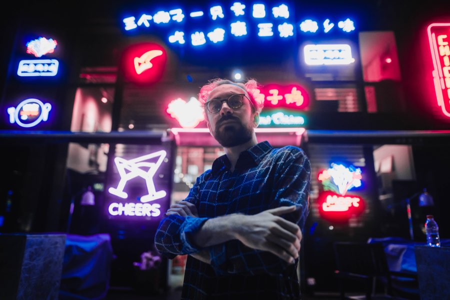 Man with arms crossed in front of colourful neon signs at night