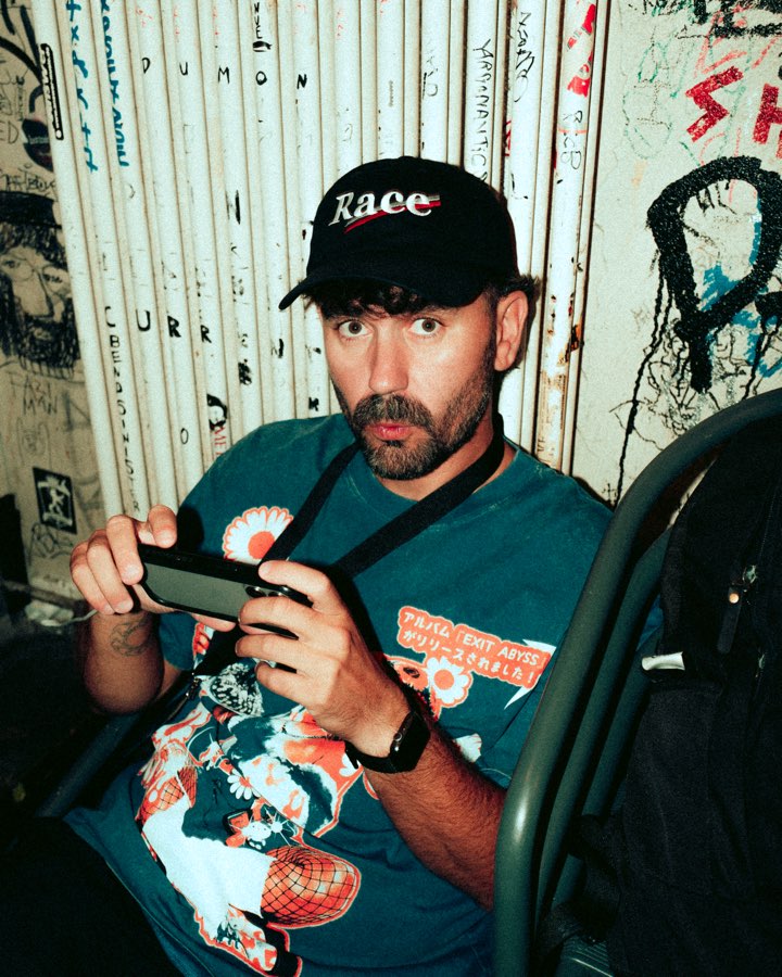 Man in a cap sitting backstage holding a phone, walls covered in stickers
