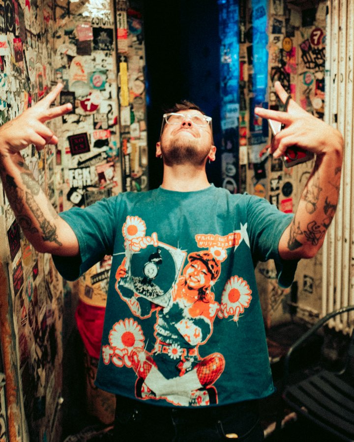 Man in sunglasses pointing upwards in a sticker-covered backstage corridor