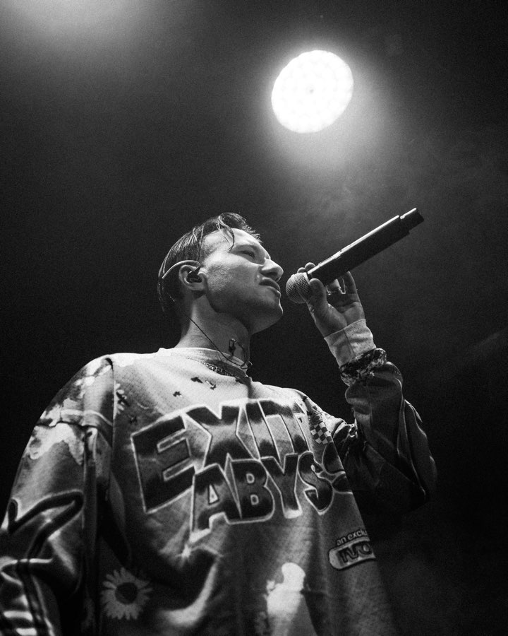 Black and white photo of a performer in an Exit Abyss jersey holding up a microphone under a spotlight