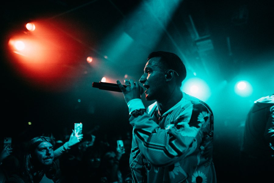 Performer in a printed shirt rapping into a microphone in teal and red light, fans filming
