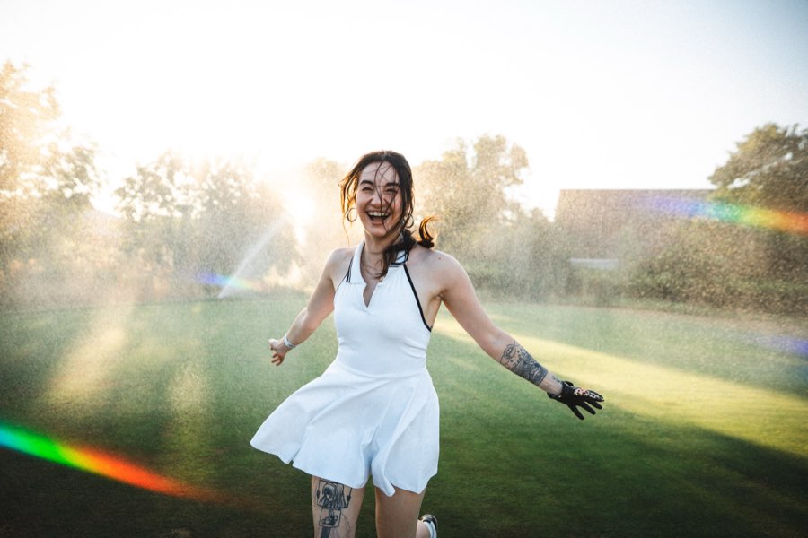 Woman in a white dress dancing through sprinkler mist with a rainbow flare