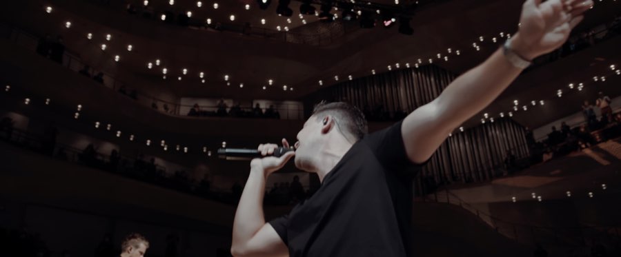 Performer singing into a microphone with his arm raised in the Elbphilharmonie concert hall