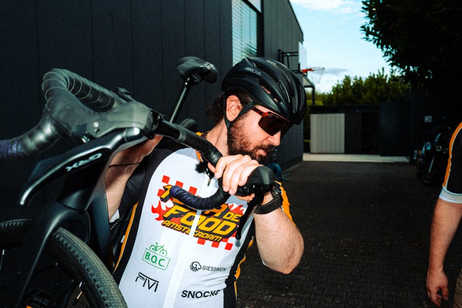 Bearded cyclist in a helmet and Tour de Food jersey carrying his bike over his shoulder