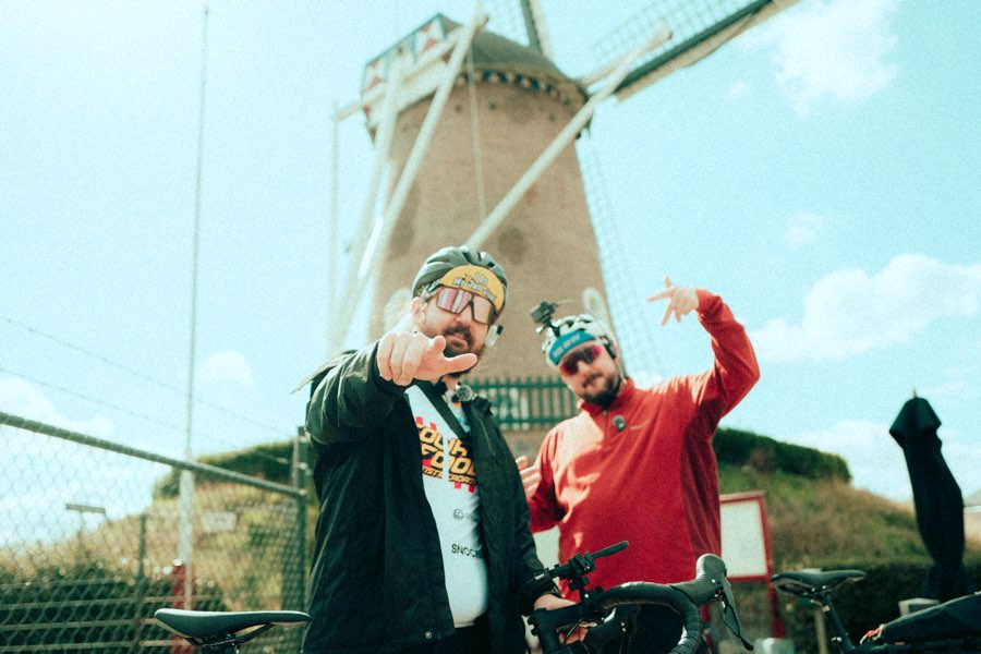 Two men in cycling gear posing in front of a windmill